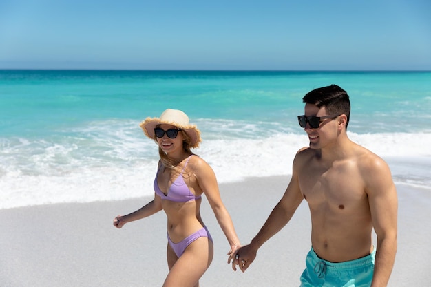 Vista frontal de um casal caucasiano caminhando na praia com o céu azul e o mar ao fundo, de mãos dadas e sorrindo