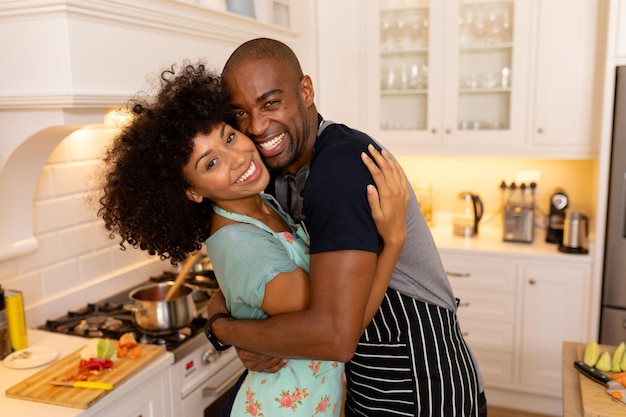 Foto vista frontal de um casal mestiço usando aventais em pé na cozinha, abraçando e sorrindo para a câmera. atividades de fim de semana em casa e estilo de vida.