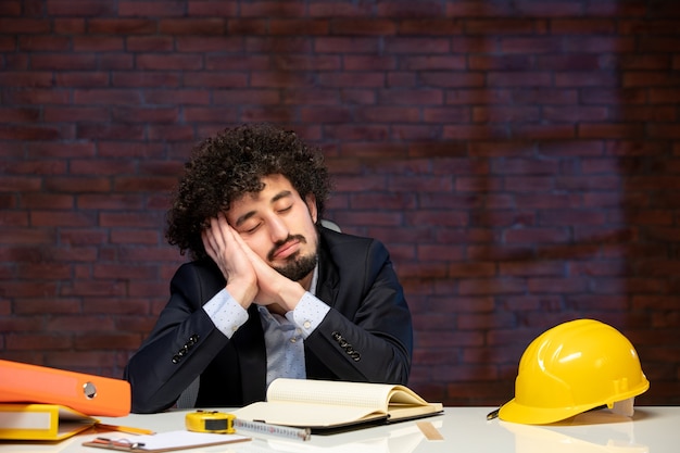 Vista frontal de um engenheiro masculino cansado sentado atrás do local de trabalho em terno empreiteiro construtor plano de ocupação de trabalho trabalho de negócios corporativos