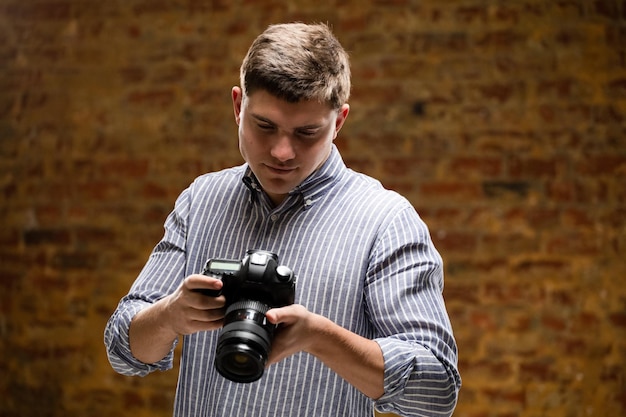 Vista frontal de um fotógrafo masculino caucasiano tirando fotos com uma câmera SLR digital em um estúdio fotográfico e verificando as imagens na tela da câmera com uma parede de tijolos ao fundo.