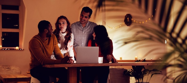 Vista frontal de um grupo de quatro jovens amigos multiétnicos, homens e mulheres, olhando juntos para um laptop e conversando na sala de estar de um apartamento à noite