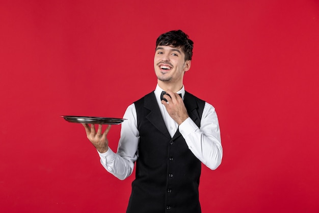 Vista frontal de um jovem garçom sorridente com um uniforme amarrando uma borboleta no pescoço segurando a bandeja na superfície vermelha isolada