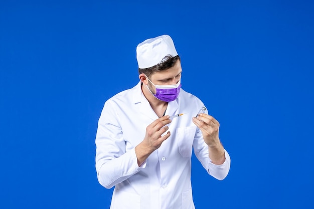 Vista frontal de um médico em traje médico e máscara segurando vacina e injeção em azul