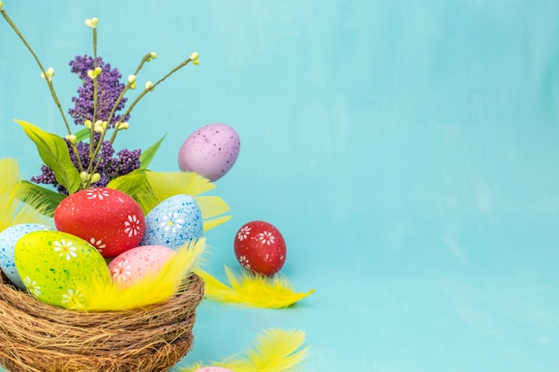 Vista frontal de um ovos de Páscoa coloridos no ninho com flores da primavera e penas amarelas sobre fundo turquesa com espaço para mensagem