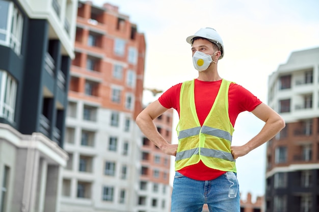 Vista frontal de um trabalhador da construção civil com uma máscara protetora e um colete reflexivo em pé entre os novos edifícios