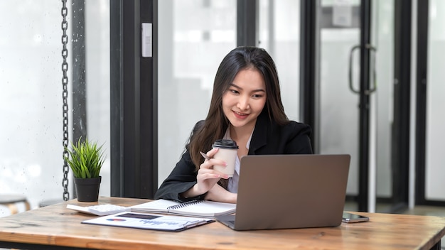 Vista frontal de uma empresária asiática segurando uma xícara de café, olhando para um documento gráfico de laptop colocado na mesa do escritório