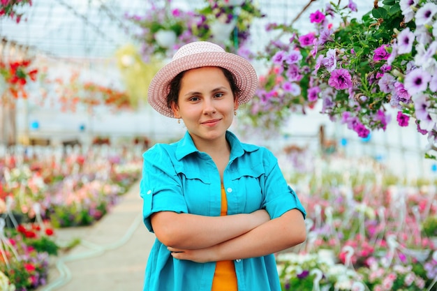 Vista frontal de uma jovem agrônoma em uma estufa a mulher cruza os braços sobre o peito e sorri brincalhão a agrônoma está de pé com um chapéu e uma camisa entre as flores