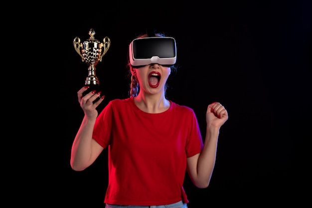 Vista frontal de uma jovem mulher jogando vr com a taça vencedora na parede escura