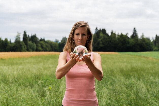 Vista frontal de uma jovem mulher meditando na natureza, segurando uma bola de cristal nas mãos com um prado e uma floresta no fundo.