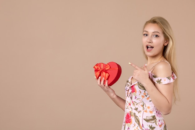 Foto vista frontal de uma jovem posando com um coração vermelho em forma de presente na parede marrom