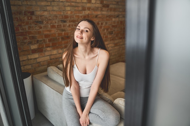 Vista frontal de uma jovem sorridente e esguia relaxando em um sofá em uma sala