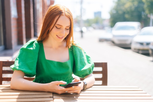 Vista frontal de uma jovem sorridente feliz usando o telefone móvel digitando, sentado à mesa no terraço do café ao ar livre em um dia ensolarado de verão. Bela ruiva apontando o dedo na tela do smartphone.