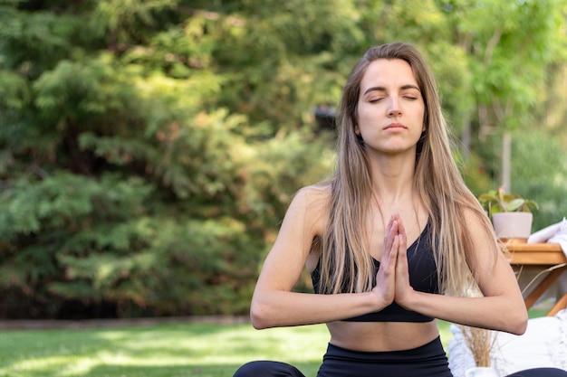 Vista frontal de uma loira meio cortada meditando com as mãos juntas e vista para o jardim atrás dela