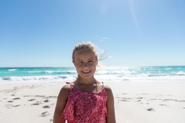 Vista frontal de uma menina caucasiana de pé na praia com o céu azul e o mar ao fundo, sorrindo para a câmera