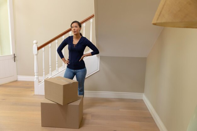 Foto vista frontal de uma mulher mestiça chegando em sua nova casa, parada no corredor olhando em volta com as mãos nos quadris, caixas de papelão no chão à sua frente