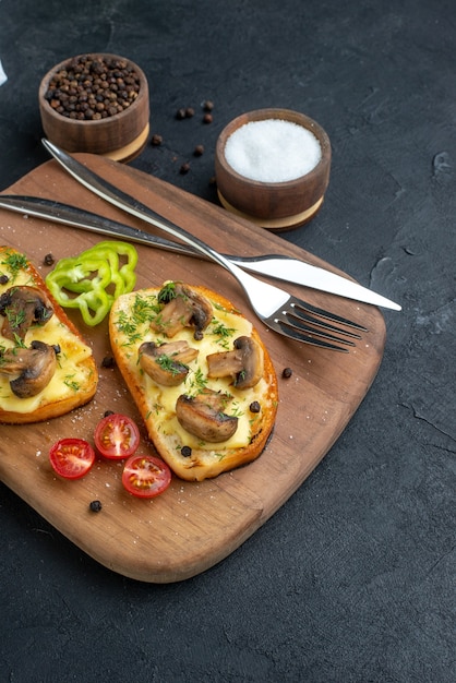 Vista frontal de deliciosos bocadillos con verduras frescas de setas y cubiertos en tabla de cortar de madera especias sobre fondo negro