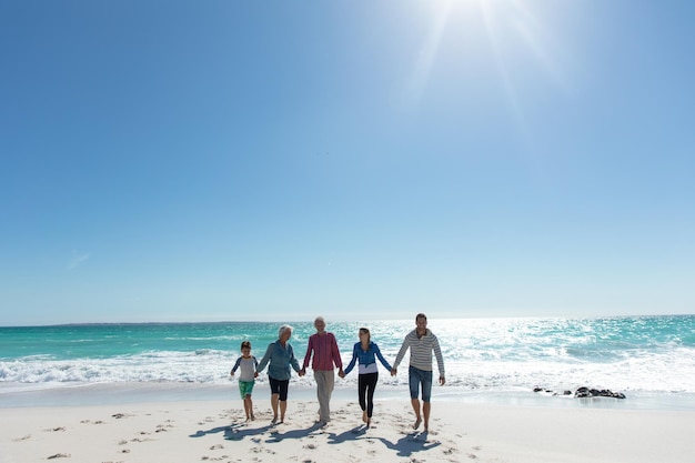 Vista frontal distante de uma família caucasiana de várias gerações na praia com o céu azul e o mar ao fundo, caminhando e de mãos dadas