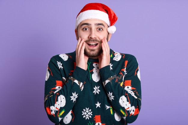 Vista frontal do cara barbudo veste o chapéu de papai noel e a camisola de nádegas com boneco de neve, olhando surpreso, mantendo as mãos perto da boca