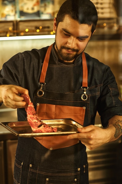 Vista frontal do chef cozinhando carne na preparação profissional da cozinha do restaurante moderno de bife antes de grelhar o belo chef barbudo brutal com tatuagem usando avental