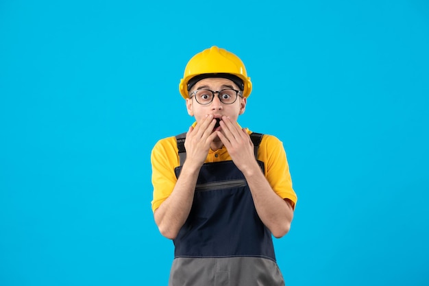 Vista frontal do construtor masculino chocado em uniforme amarelo sobre azul