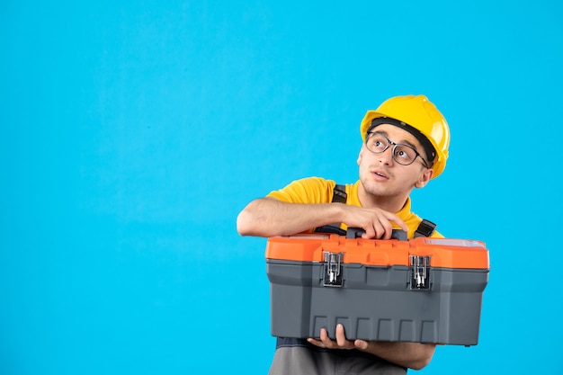 Vista frontal do construtor masculino de uniforme com caixa de ferramentas nas mãos na parede azul