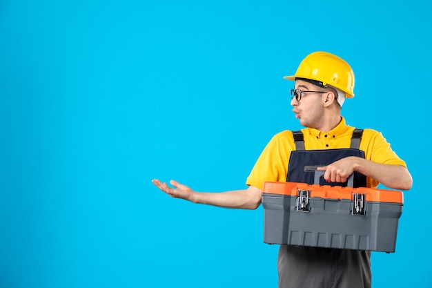 Vista frontal do construtor masculino de uniforme e capacete com caixa de ferramentas na parede azul