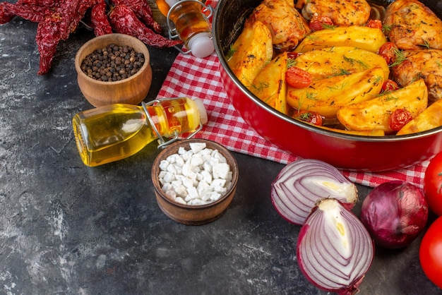 Vista frontal do delicioso jantar com frangos batatas verdes em panela na toalha despojada vermelha dobrada pimentas secas garrafas de óleo caídas legumes