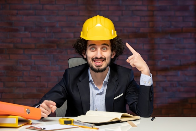 vista frontal do engenheiro masculino sentado atrás do local de trabalho em terno e capacete amarelo plano de negócios construtor ocupação trabalho corporativo projeto de trabalho