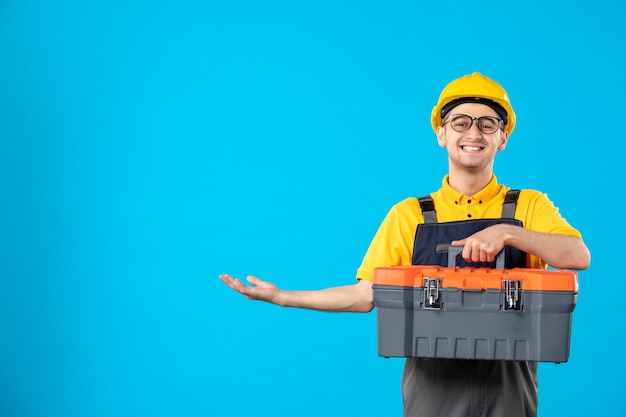 Vista frontal do feliz construtor masculino de uniforme e capacete com caixa de ferramentas na parede azul