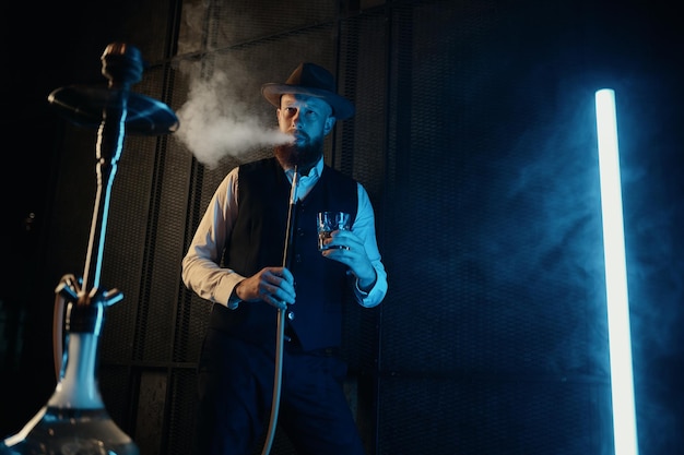 Vista frontal do homem descansando fumando cachimbo de água no local de trabalho em casa. Cara retrô bebendo chapéu relaxa no quarto escuro com iluminação neon