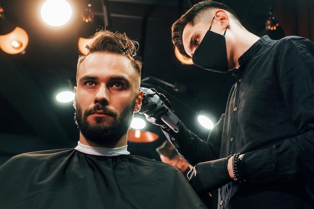 Vista frontal do jovem barbudo que está sentado e cortando o cabelo na barbearia por cara com máscara protetora preta