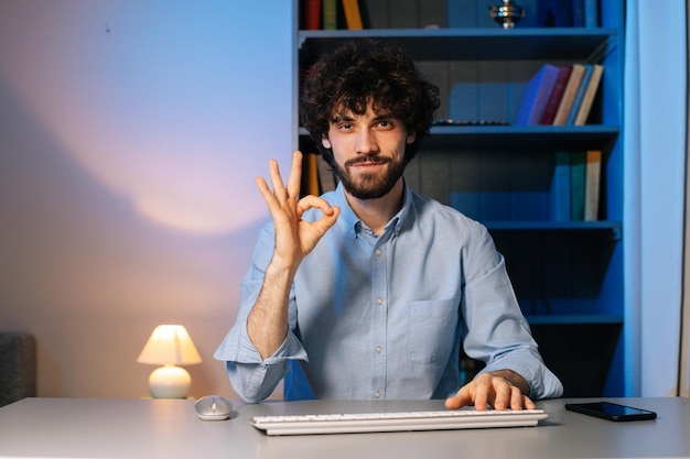 Vista frontal do jovem barbudo sorridente mostrando o gesto de OK olhando para a câmera durante o trabalho no computador no escritório em casa. Conceito de trabalho remoto. Conceito de trabalho remoto.