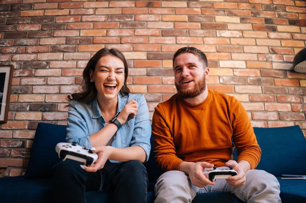 Vista frontal do jovem casal feliz rindo segurando controladores e jogando videogame no console sentados juntos no sofá