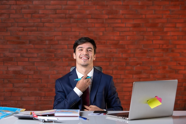 vista frontal do jovem empresário sorridente sentado atrás de seu local de trabalho com o laptop em um terno plano de trabalho empreiteiro ocupação corporativa construtor projeto negócios trabalho