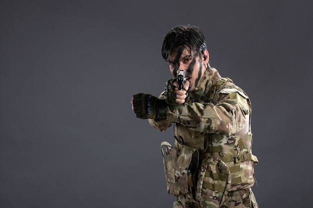 Vista frontal do jovem soldado camuflado com arma na parede escura