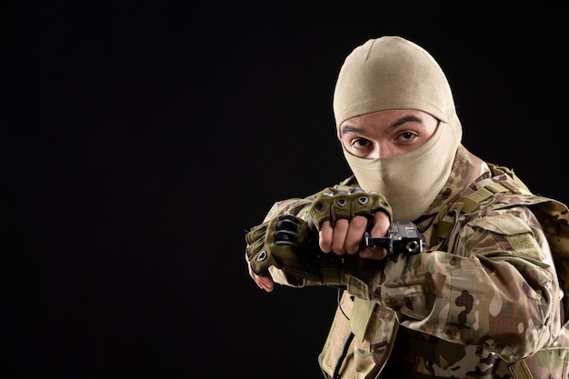 Foto vista frontal do jovem soldado de uniforme apontando a arma na parede preta