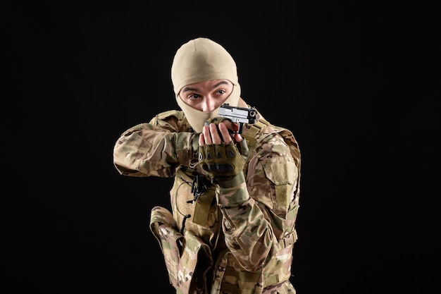 Vista frontal do jovem soldado de uniforme apontando a arma na parede preta