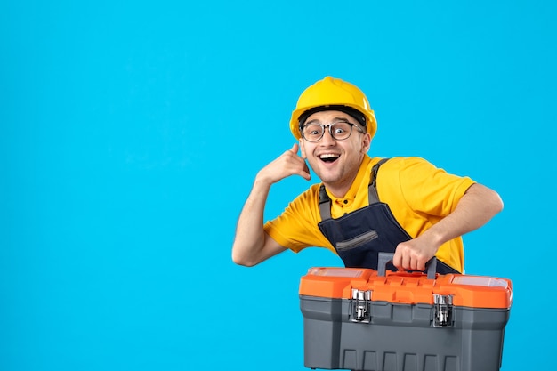 Vista frontal do trabalhador apressado em uniforme amarelo carregando caixa de ferramentas na parede azul