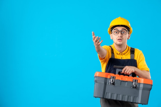 Vista frontal do trabalhador do sexo masculino em uniforme amarelo carregando caixa de ferramentas no azul