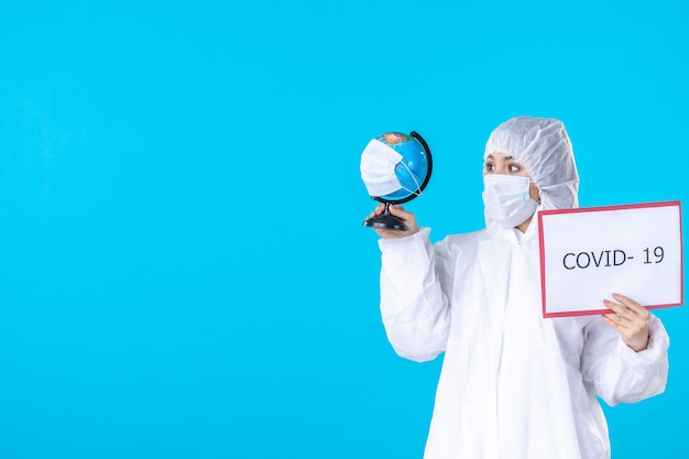 Foto vista frontal doctora en traje de protección y máscara sobre fondo azul covid- aislamiento de salud pandemia de virus de ciencia médica globe