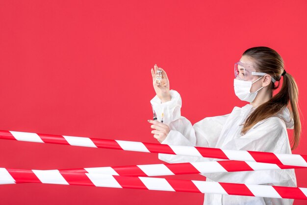 Vista frontal doctora en traje de protección sosteniendo inyecciones sobre fondo rojo enfermera covid- máscara de salud hospital curar tejido virus emoción