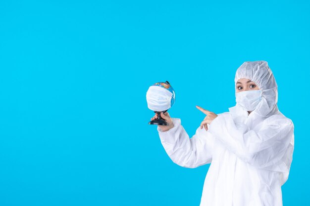 Vista frontal doctora en traje protector y máscara sosteniendo globo enmascarado sobre el fondo azul ciencia color covid- virus pandémico médico