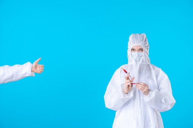 Vista frontal doctora en traje protector y máscara sosteniendo la inyección sobre el fondo azul hospital médico virus pandémico covid ciencia de la salud