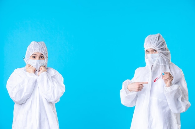 Vista frontal doctoras en trajes protectores y máscaras sosteniendo frascos sobre fondo azul salud hospitalaria covid- virus pandémico médico