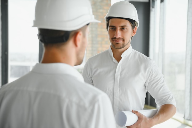 Una vista frontal de dos arquitectos inteligentes con cascos blancos revisando planos en un sitio de construcción en un día soleado