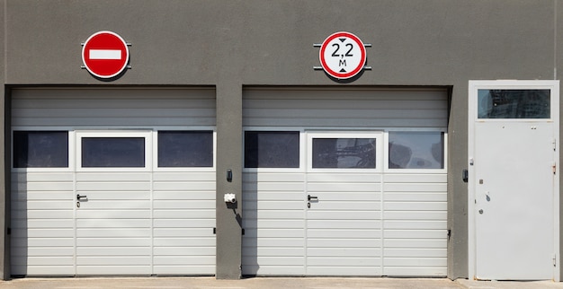 Vista frontal en dos puertas de garaje blancas cerradas y puerta de entrada electrónica, paredes de molduras grises y señales de tráfico.