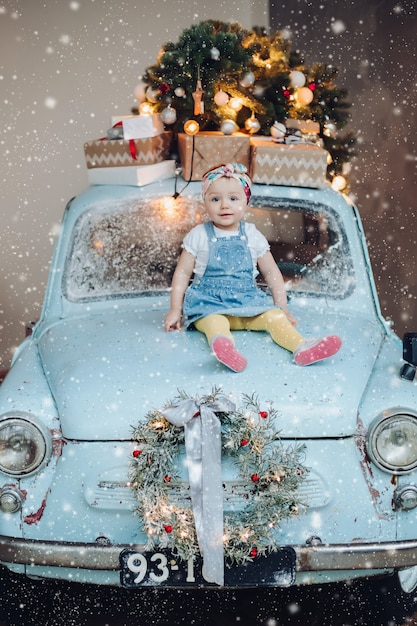 Vista frontal de dulce y de moda linda niña sentada en el coche retro azul decorado para Navidad.