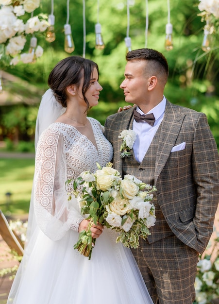 Vista frontal del elegante novio manteniendo las manos en el bolsillo mirando a la novia que se vistió con un vestido con elementos étnicos sosteniendo