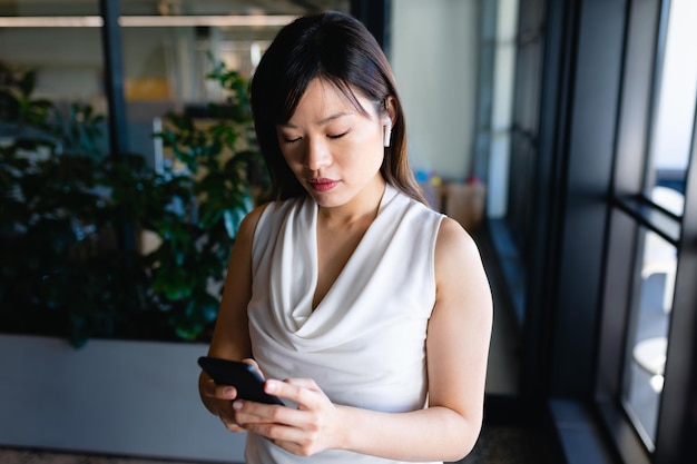 Vista frontal de una empresaria asiática vestida con ropa elegante y auriculares, trabajando en la oficina moderna, parada junto a una ventana y usando su teléfono inteligente.