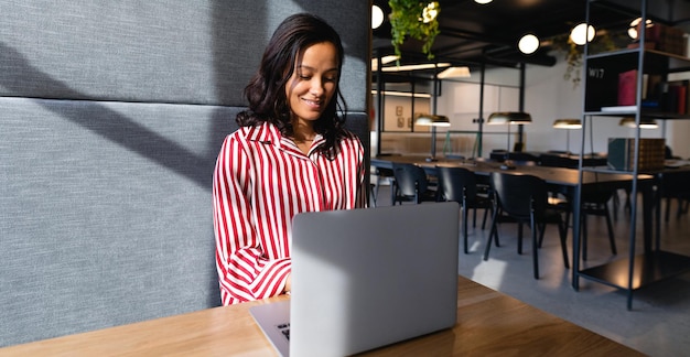 Vista frontal de una empresaria de raza mixta que trabaja en una oficina moderna, sentada junto a un escritorio y usando una computadora portátil. Distanciamiento social y autoaislamiento en cuarentena